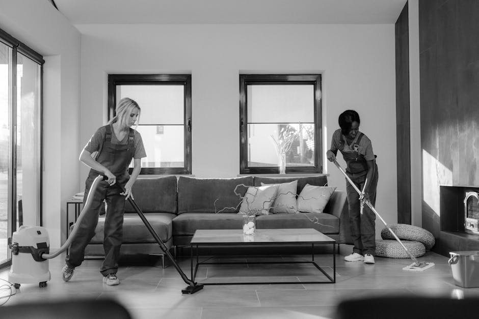 A woman performing surface cleaning on a tall, dark-colored tiled wall in a bright room with two large windows, using an extendable cleaning tool. She is wearing casual work clothes and is focused on