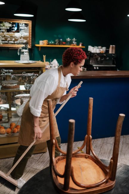 A cleaner performing surface cleaning outside a commercial art store, with a squeegee or cloth in hand, focused on maintaining hygiene and cleanliness. The store's glass window features white text in 